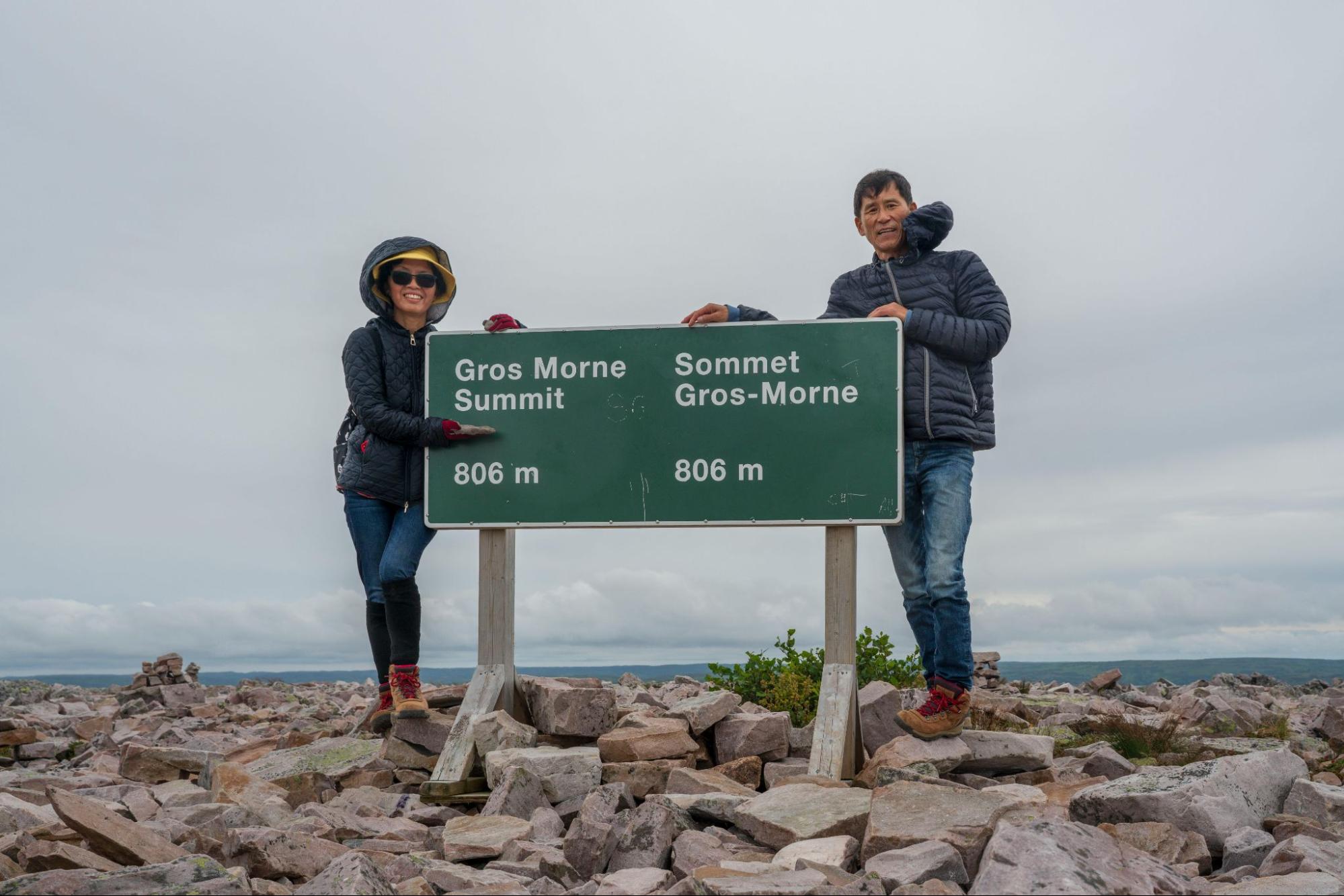 A woman and a man stand on a cliff near a sign for the Gros Morne Mountain